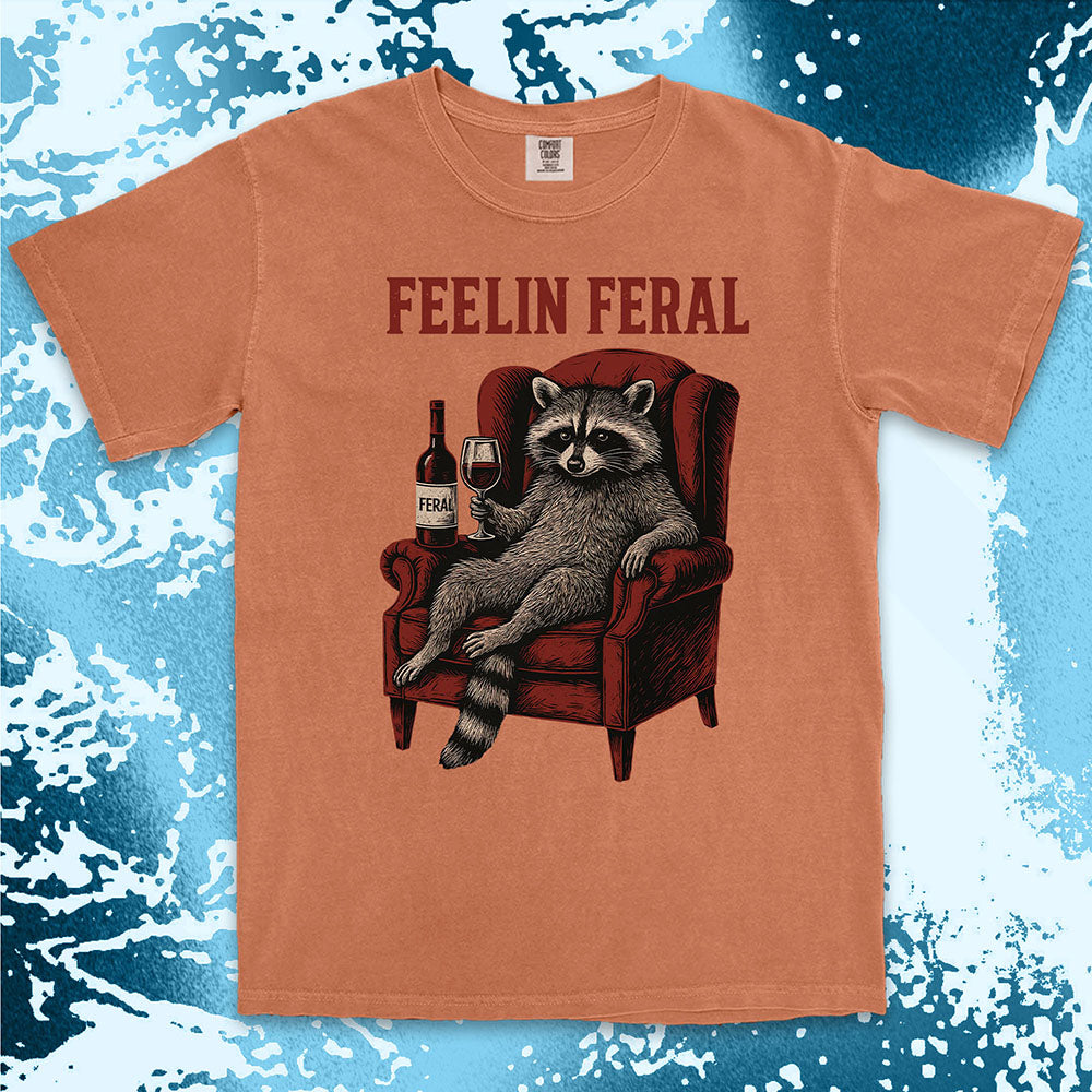 Orange t-shirt with a raccoon graphic and 'FEELIN FERAL' text on a blue and white abstract background