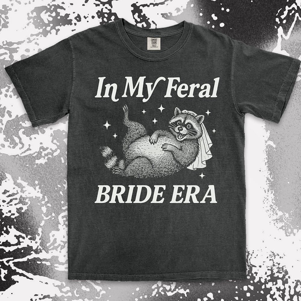 Comfort Color t-shirt with a raccoon wearing a bridal veil and text ÒIn My Feral Bride EraÓ in playful vintage lettering.