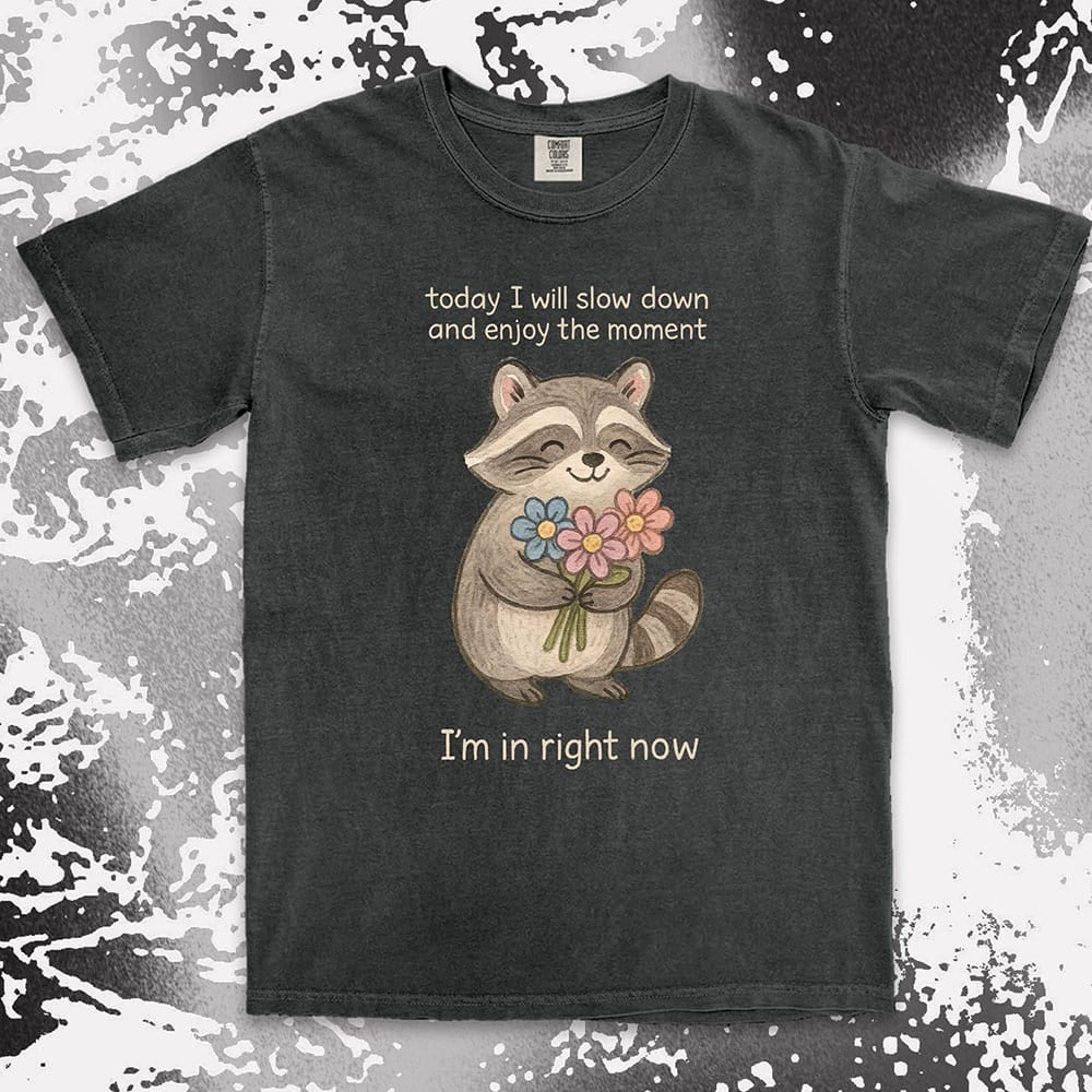Comfort Color t-shirt with a smiling raccoon holding flowers and text ÒToday I will slow down and enjoy the moment Ð IÕm in right nowÓ in a soft, mindful design.