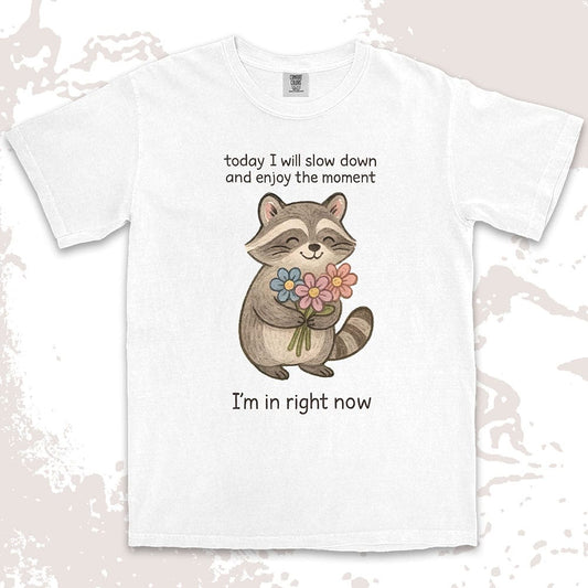 Comfort Color t-shirt with a smiling raccoon holding flowers and text ÒToday I will slow down and enjoy the moment Ð IÕm in right nowÓ in a soft, mindful design.