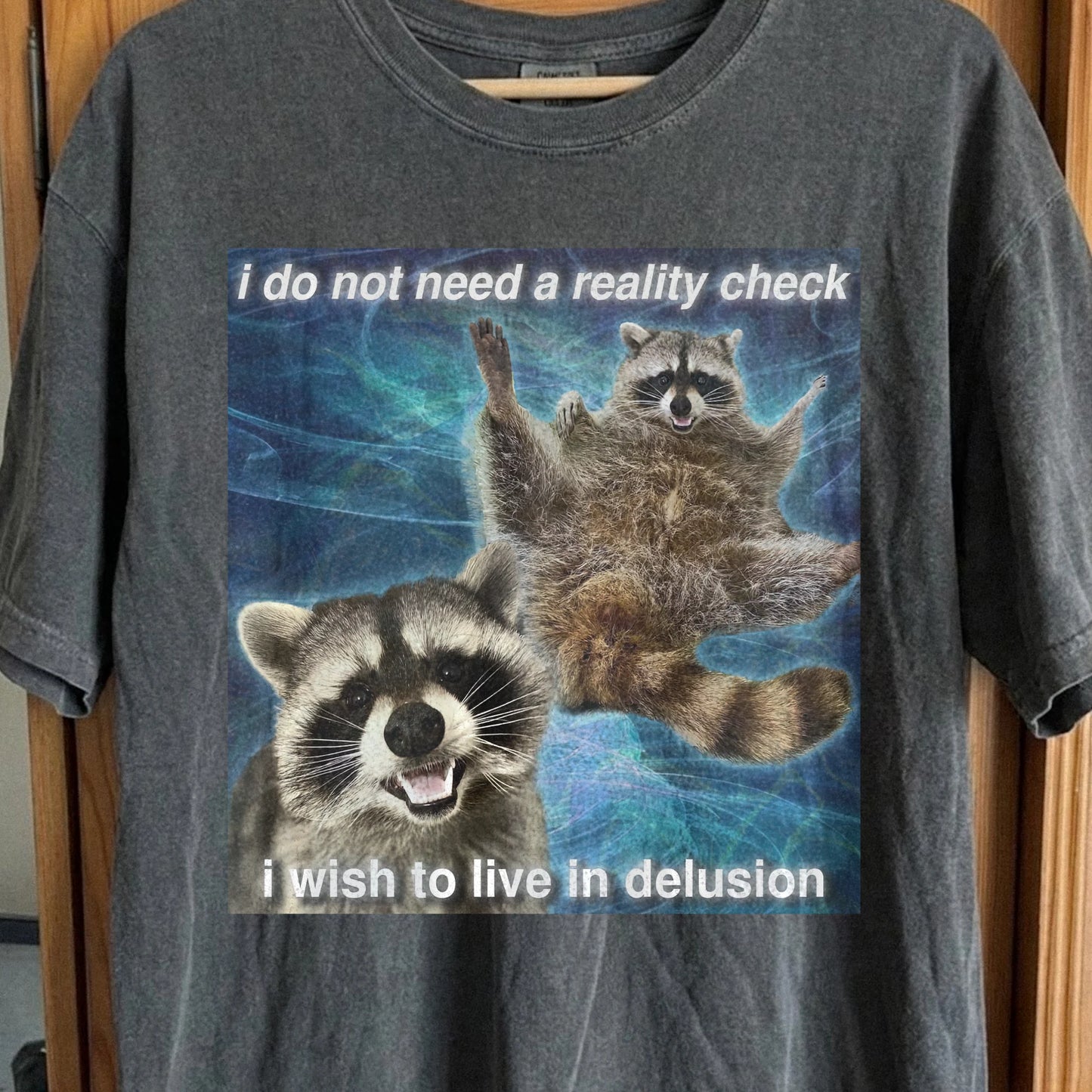 Raccoon t-shirt with quote “I do not need a reality check, I wish to live in delusion” on surreal blue background