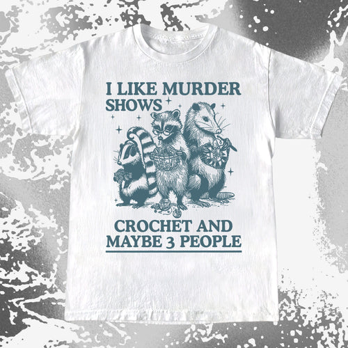 White t-shirt with raccoon graphic and text 'I like murder shows, crochet and maybe 3 people' on a textured background.