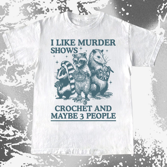 White t-shirt with raccoon graphic and text 'I like murder shows, crochet and maybe 3 people' on a textured background.