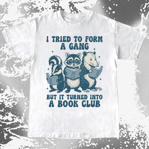 White t-shirt with cartoon raccoons and opossum reading books and text on a textured gray background