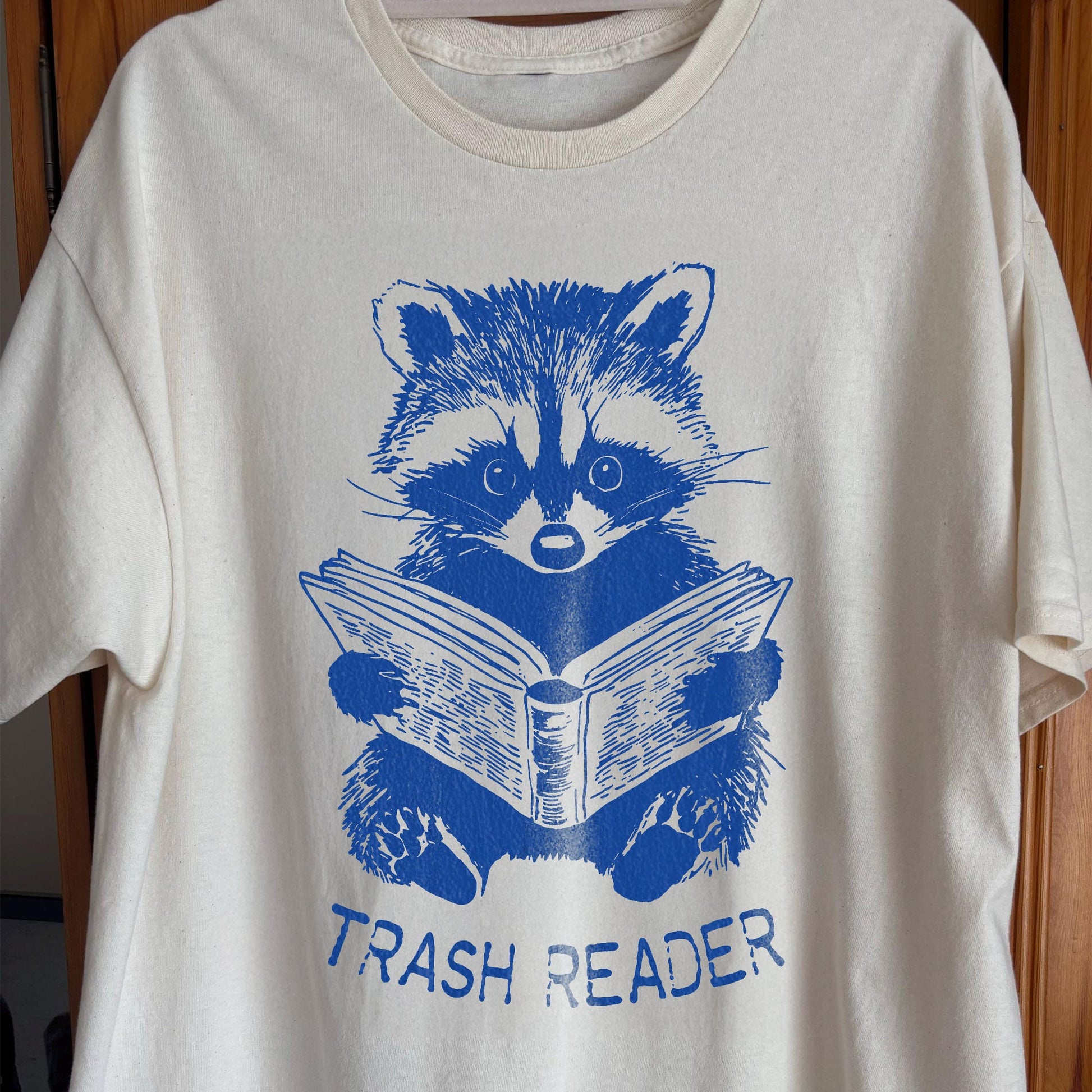 Blue raccoon illustration reading a book with the text Trash Reader in a funny book lover design