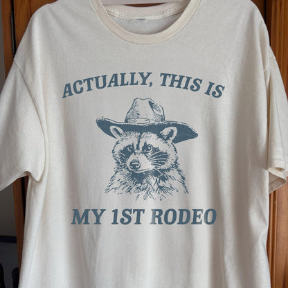 White t-shirt with a graphic of a raccoon wearing a hat and text 'Actually, this is my 1st Rodeo' on a wooden background.