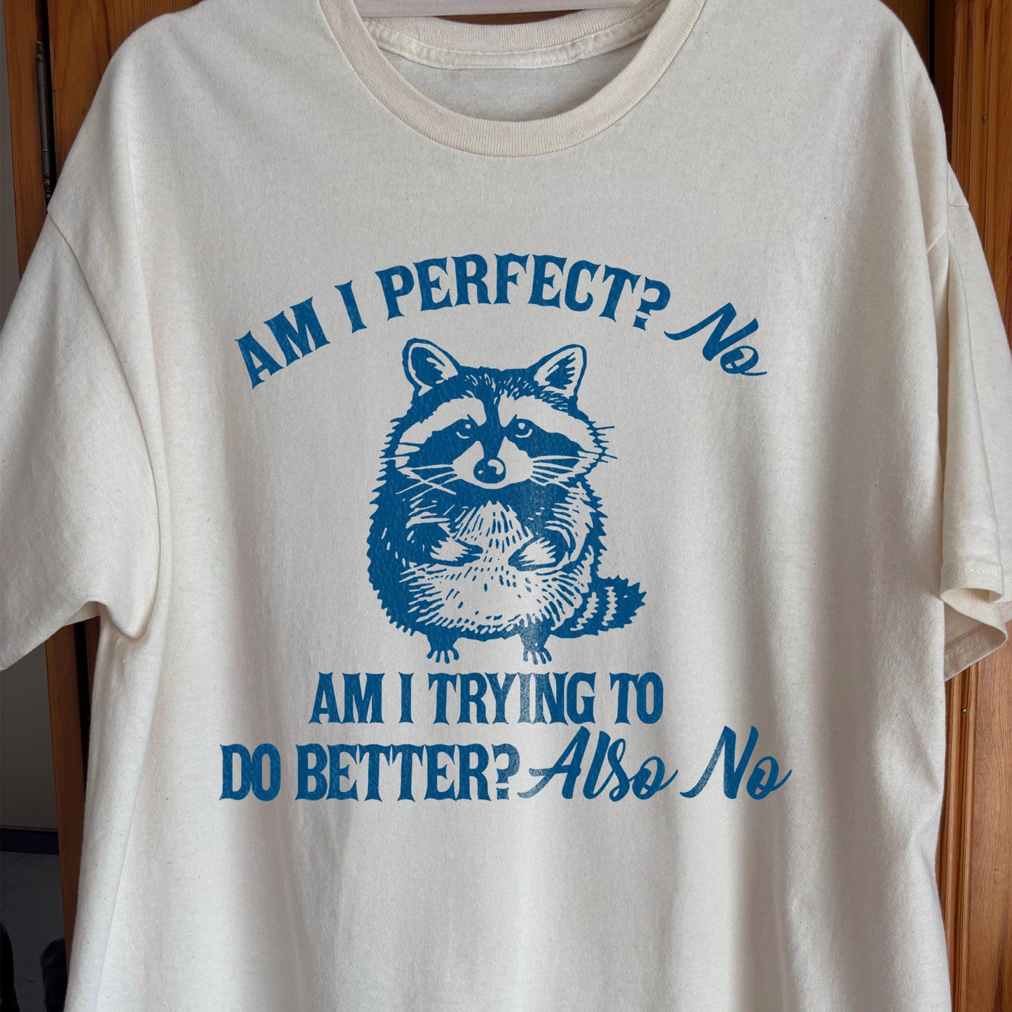 Raccoon graphic t-shirt with quote “Am I perfect? No. Am I trying to do better? Also no” in blue ink