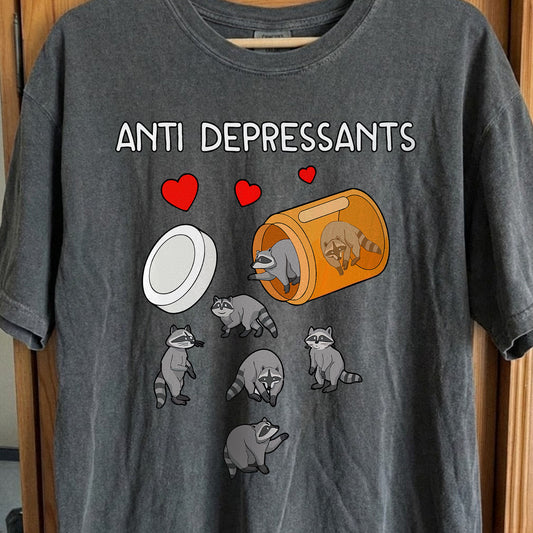 Gray t-shirt with cartoon raccoons and 'ANTI DEPRESSANTS' text on a wooden surface