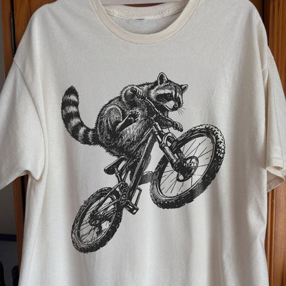 White t-shirt with a black and white illustration of a raccoon riding a bicycle on a wooden surface.