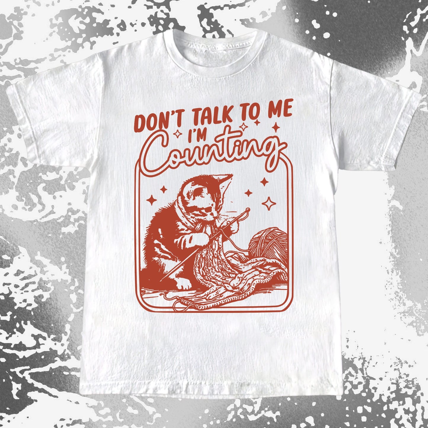 White t-shirt with red graphic of a cat counting and text 'Don't talk to me I'm counting' on a textured gray background.