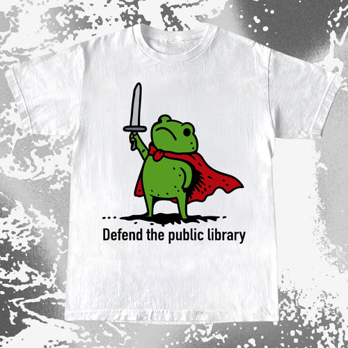 White t-shirt with a green superhero frog graphic and text 'Defend the public library' on a textured gray background.