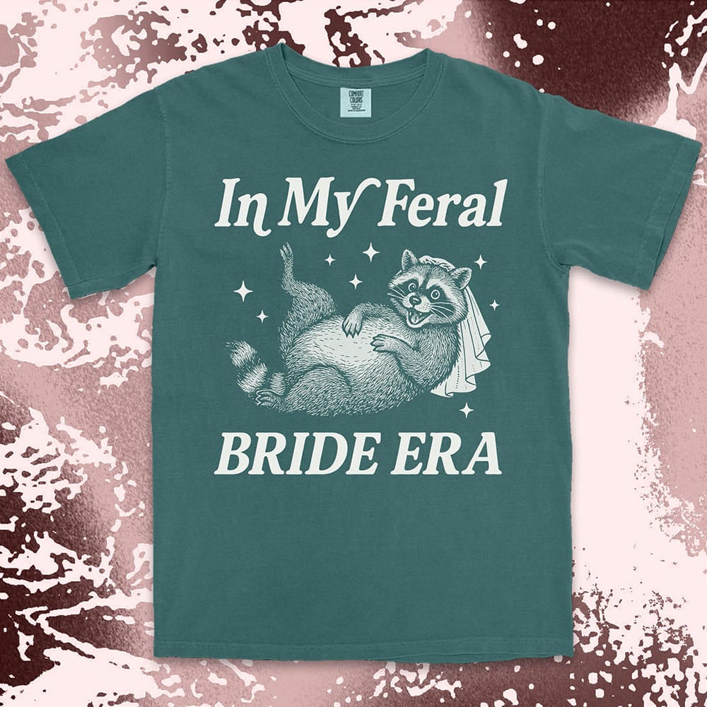 Comfort Color t-shirt with a raccoon wearing a bridal veil and text ÒIn My Feral Bride EraÓ in playful vintage lettering.