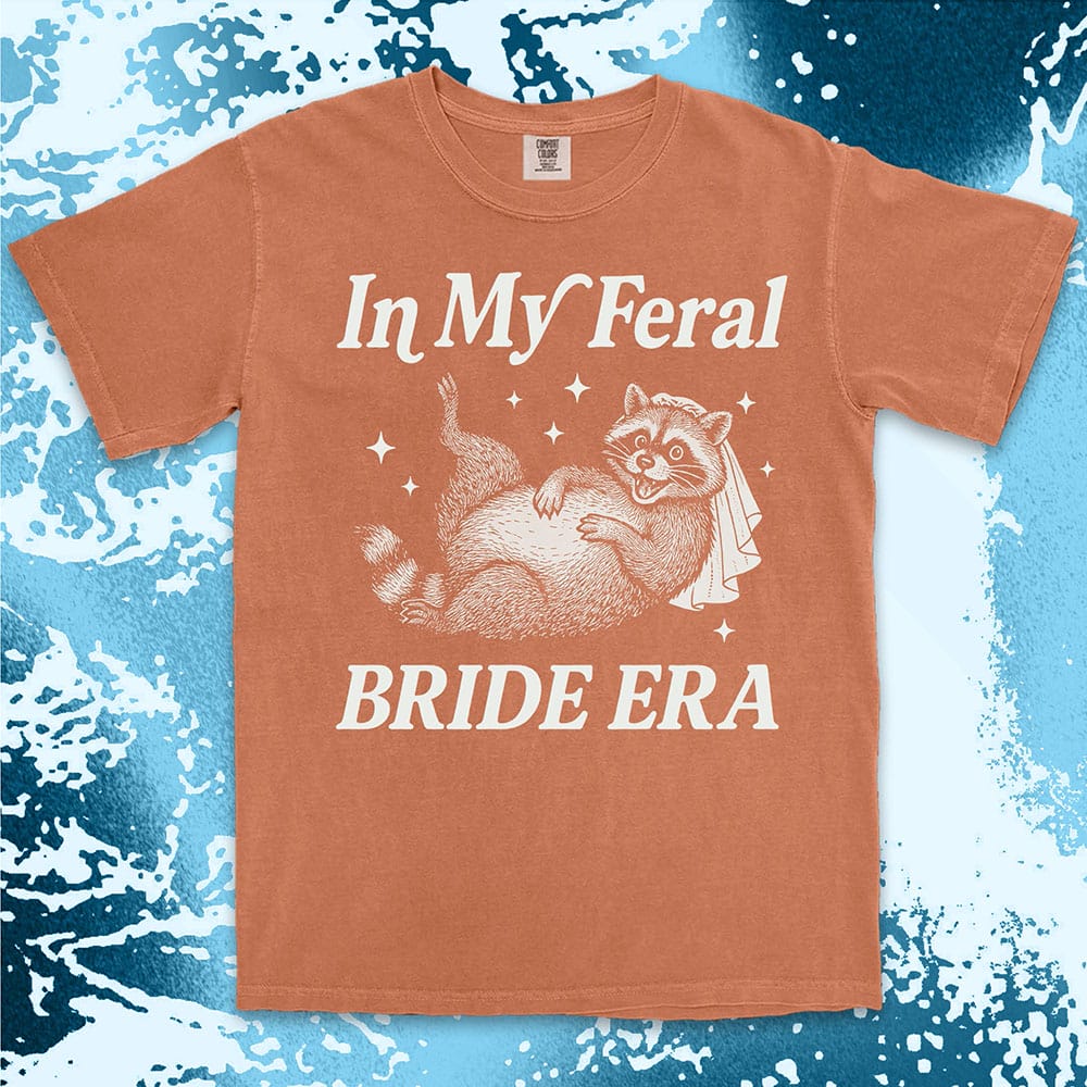 Comfort Color t-shirt with a raccoon wearing a bridal veil and text ÒIn My Feral Bride EraÓ in playful vintage lettering.