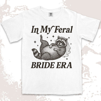 Comfort Color t-shirt with a raccoon wearing a bridal veil and text ÒIn My Feral Bride EraÓ in playful vintage lettering.