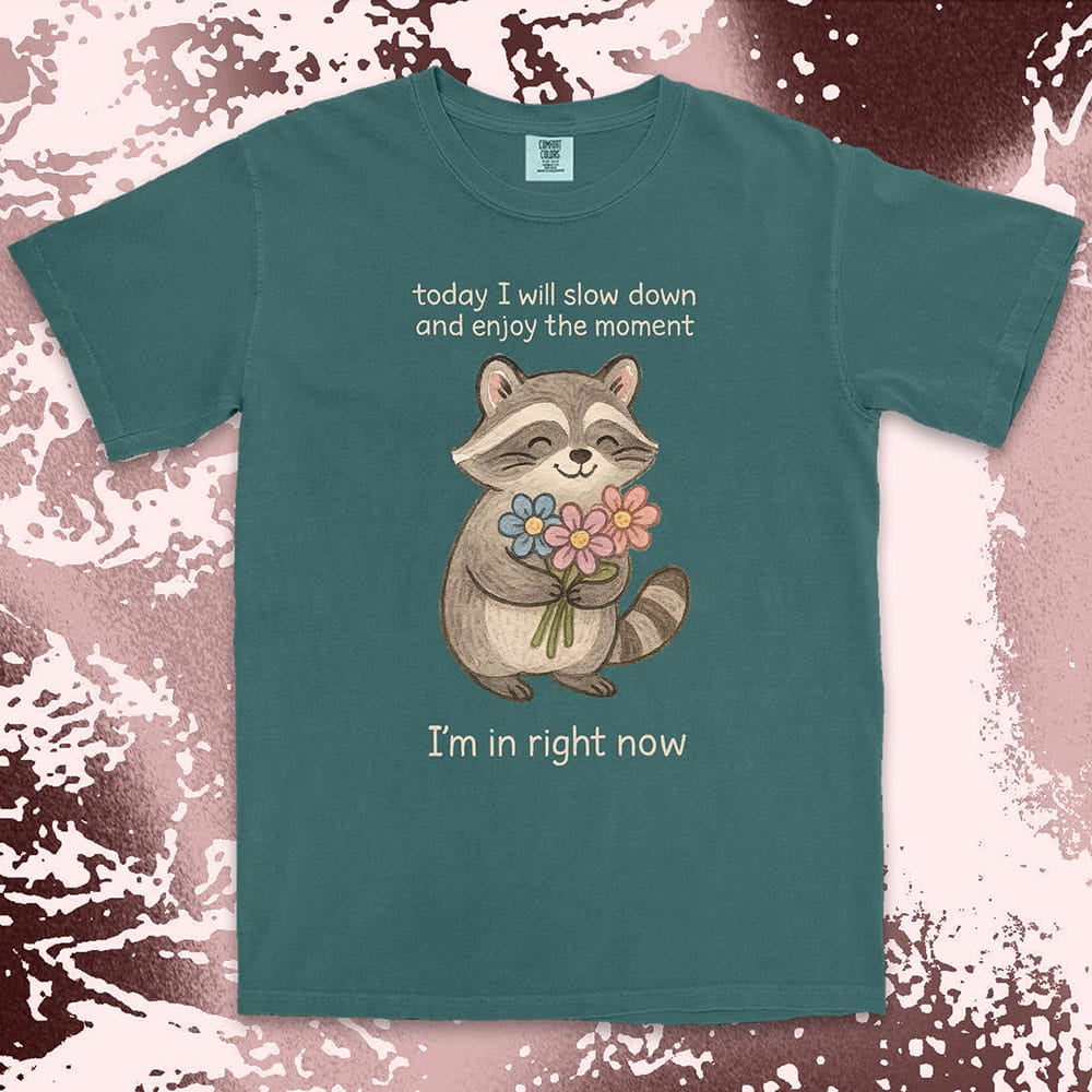 Comfort Color t-shirt with a smiling raccoon holding flowers and text ÒToday I will slow down and enjoy the moment Ð IÕm in right nowÓ in a soft, mindful design.