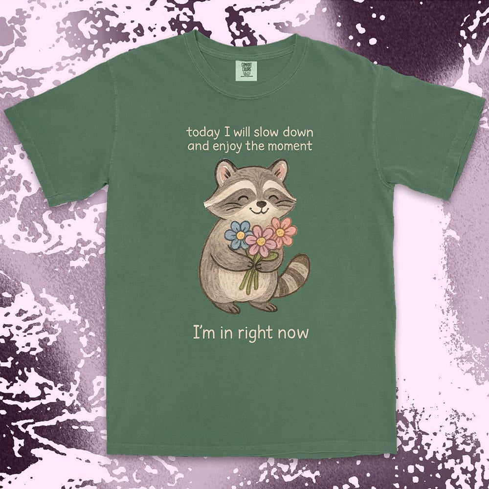 Comfort Color t-shirt with a smiling raccoon holding flowers and text ÒToday I will slow down and enjoy the moment Ð IÕm in right nowÓ in a soft, mindful design.