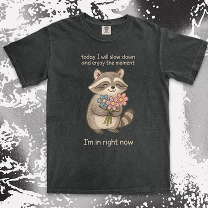 Comfort Color t-shirt with a smiling raccoon holding flowers and text ÒToday I will slow down and enjoy the moment Ð IÕm in right nowÓ in a soft, mindful design.