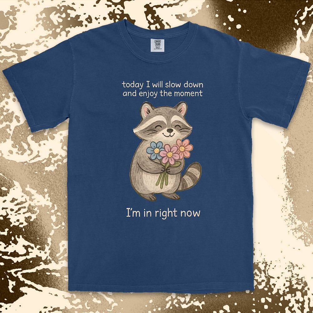 Comfort Color t-shirt with a smiling raccoon holding flowers and text ÒToday I will slow down and enjoy the moment Ð IÕm in right nowÓ in a soft, mindful design.