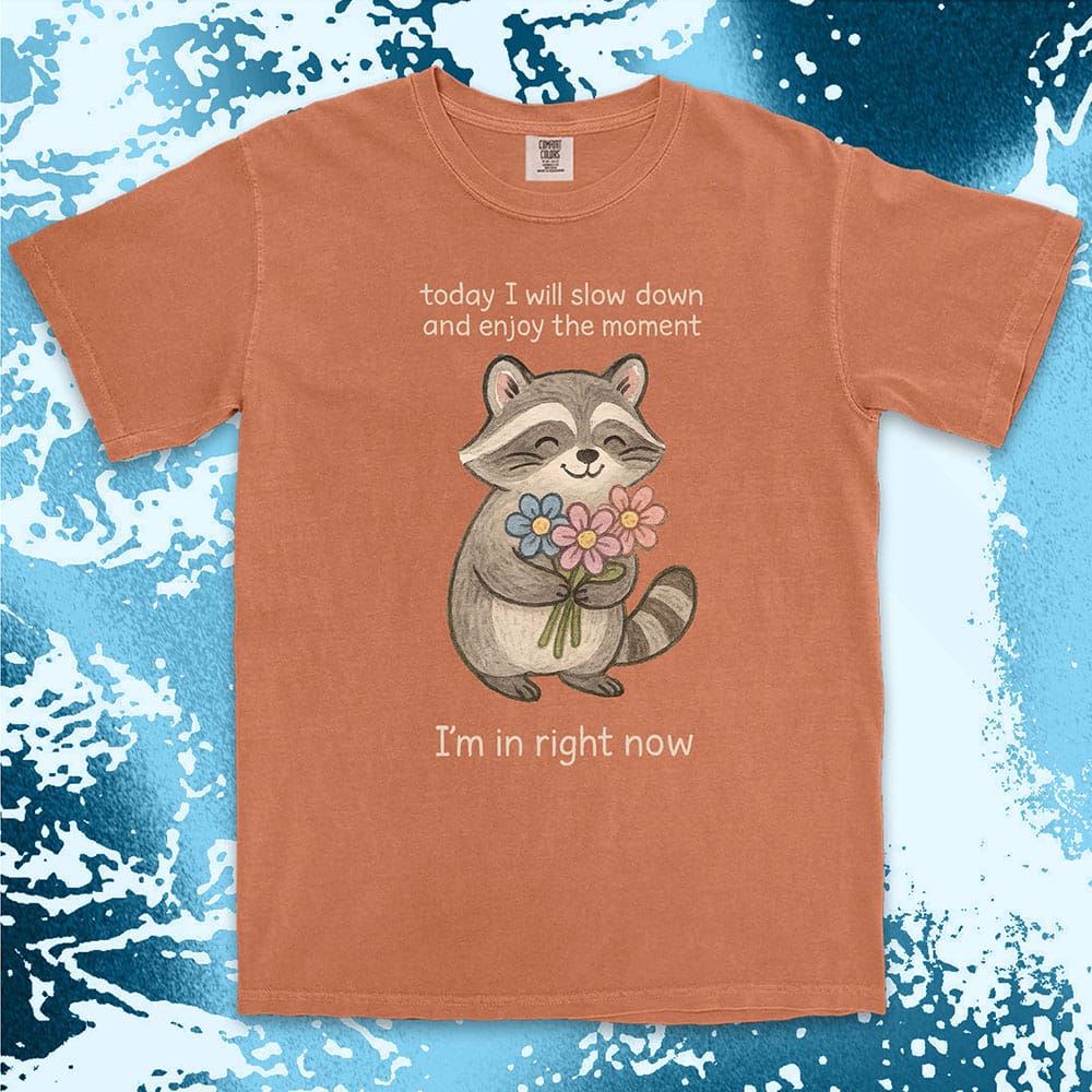 Comfort Color t-shirt with a smiling raccoon holding flowers and text ÒToday I will slow down and enjoy the moment Ð IÕm in right nowÓ in a soft, mindful design.