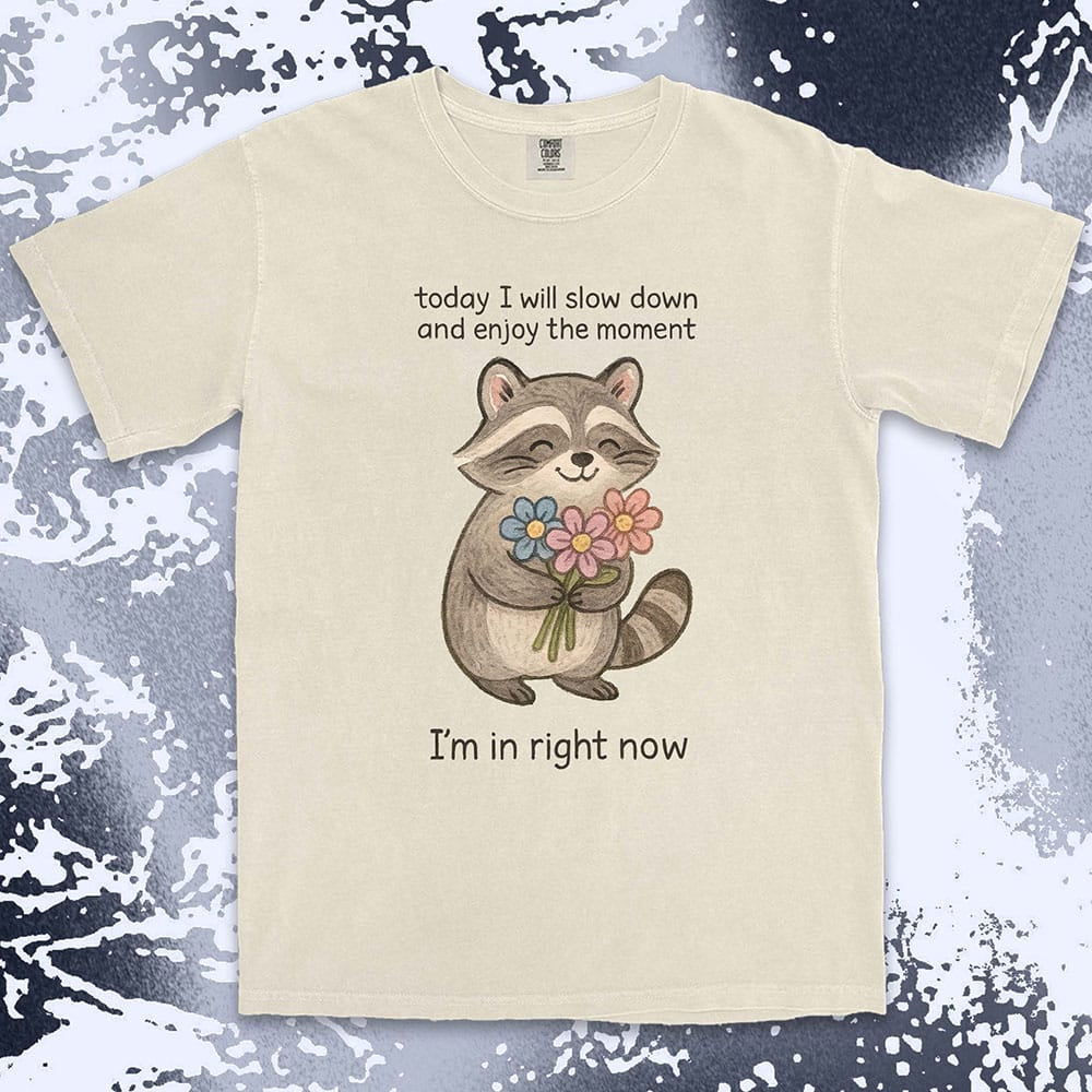 Comfort Color t-shirt with a smiling raccoon holding flowers and text ÒToday I will slow down and enjoy the moment Ð IÕm in right nowÓ in a soft, mindful design.