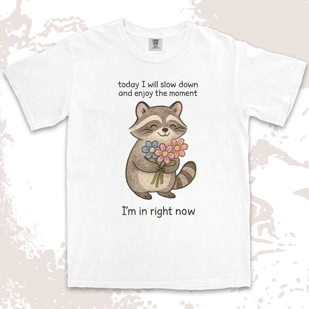 Comfort Color t-shirt with a smiling raccoon holding flowers and text ÒToday I will slow down and enjoy the moment Ð IÕm in right nowÓ in a soft, mindful design.